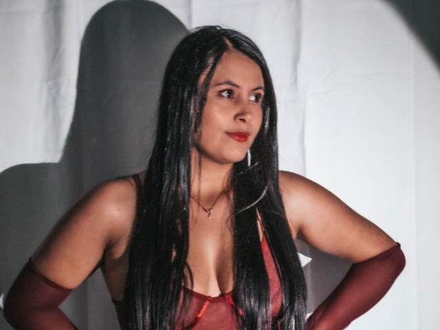 EmilySavage with long black hair, red lipstick, red dress, showing her tits, wearing red gloves, hands on hips, looking to the side, shadow on white wall.