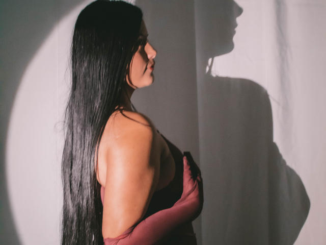 EmilySavage with long black hair, wearing a red dress and gloves, side profile, shadow on white wall, hands covering her tits, light highlighting her tan skin.