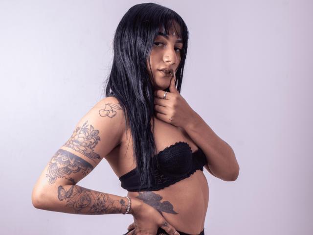 BellaHoffis with long black hair, wearing black lace bra and fishnet pants, showing tattoos on arm and side, hands in hair, looking at camera, standing in three-quarters pose.