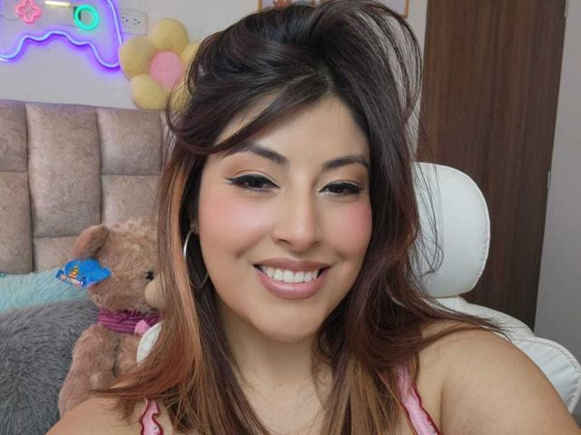 AlexaQueenOne with brown hair, pink lipstick, hoop earrings, smiling, wearing pink top, holding brown teddy with blue tag, neon wall decoration, and white chair in background.