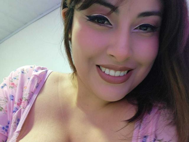 AlexaQueenOne with pink floral top, smiling, showing her tits, brown hair, pink eyeliner, and pink lipstick. Her skin is fair, and she has a cute face.
