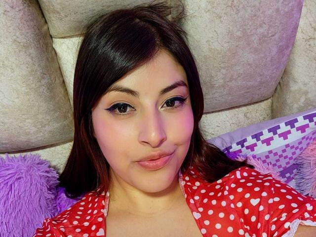 AlexaQueenOne with dark brown hair, black eyeliner, and pink lipstick, wearing a red polka dot shirt, lying on purple and white pillows, smiling slightly.