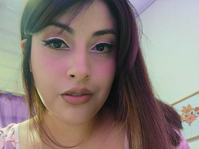 AlexaQueenOne has pink-tinted skin, dark brown eyes with thick eyelashes, and pink lipstick. She's wearing a pink floral top with straight brown hair.