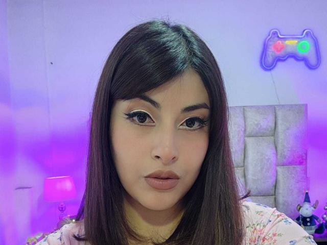 AlexaQueenOne has long brown hair, large brown eyes with thick eyelashes, and full pink lips. She's wearing a floral shirt, looking straight at the camera with a neutral expression.