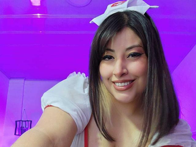 AlexaQueenOne with medium brown hair, wearing a white nurse cap and dress, smiling at the camera, purple lighting in the background, showing her cute face and small tits.