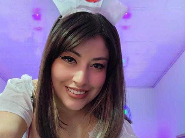 AlexaQueenOne has brown hair, white eyeliner, and is smiling with pink lips. She wears a white headband and white top, with purple and pink lights in the background.