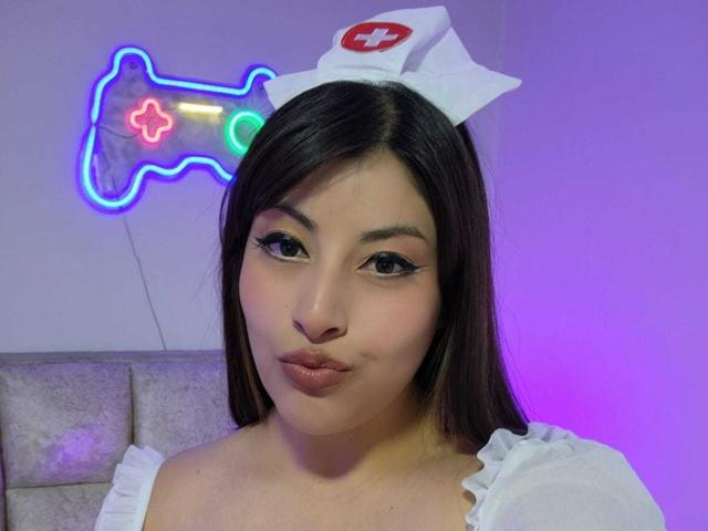 AlexaQueenOne with dark brown hair, wearing a white nurse cap and off-shoulder top, pouting her plump, glossy lips. Neon game controller light up behind her.