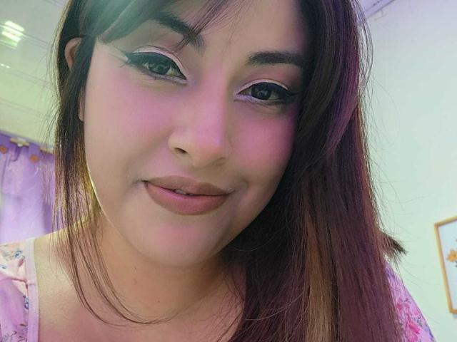 AlexaQueenOne has pinkish skin, dark brown eyes with white eyeliner, and brown hair with purple highlights. She's smiling with pink lipstick, wearing a pink floral top.