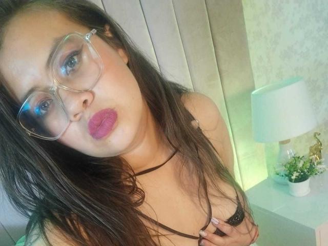 IsabellaReid with long brown hair, wearing clear glasses and purple lipstick, pouting, showing her tits with a black choker, in a bedroom with a green-lit lamp.