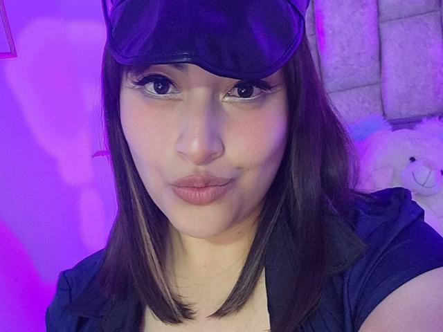 AlexaQueenOne with purple-lit face, dark brown straight hair, black visor, and pouty lips, wearing a black shirt, showing a small beauty mark on her left cheek.