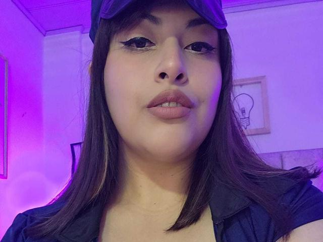 AlexaQueenOne has medium brown skin, straight brown hair, black eyeliner, and wears a purple cap with a black shirt. She's looking at the camera with slightly parted lips.