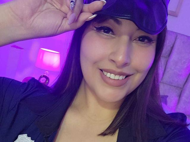 AlexaQueenOne with dark hair, wearing a black cap and shirt, smiling, showing white teeth, holding cap with purple-lit background, visible lamp, and soft textures in the room.