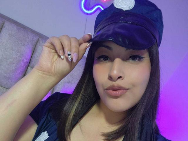 AlexaQueenOne with puffy lips, dark straight hair, wearing a police cap, touching her hat, showing black nail with white skull, purple and blue neon light in background.