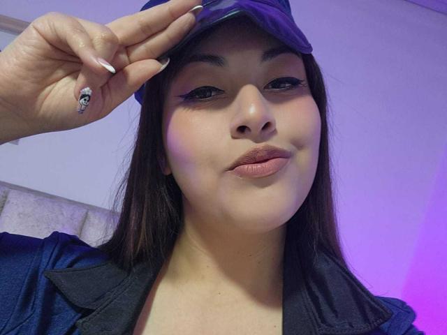 AlexaQueenOne with dark hair, black eyeliner, and pink lipstick, saluting with a skull ring, wearing a blue cap, and black jacket, showing her pouty, cute face.