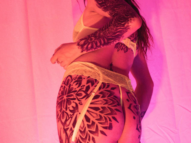 SofiFw's tattooed ass in yellow lace garter and bra, pink-tinged lighting, holding back her top to show off intricate black mandala tattoos on her buttocks and arm.