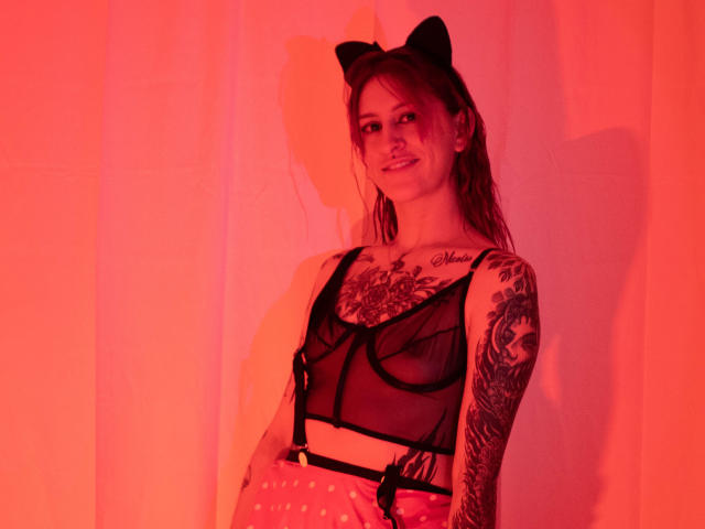 SofiFw wearing black cat ears, sheer black bra, and polka dot pants, showing tattoos on her right arm and chest, under red lighting, with a slight smile.
