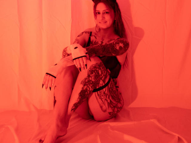 SofiFw with tattoos, red lighting, sitting on white cloth, wearing black lingerie, cat ears, and white gloves, smiling, showing her tits and pussy, legs crossed, arms hugging knees.