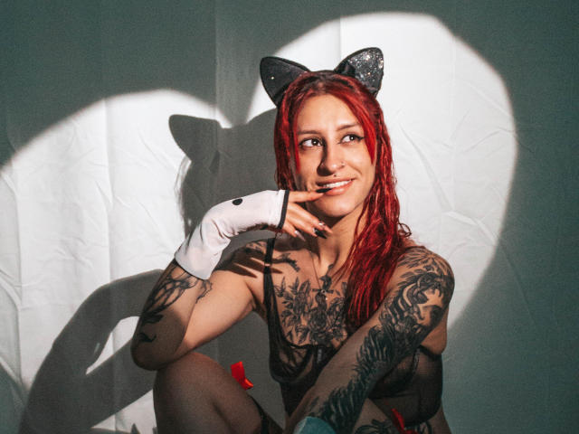 SofiFw with red hair, cat ears, and tattoos, topless, wearing white glove, finger near mouth, smiling, heart shadow, sunlight, and rose petals in photo.