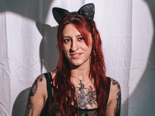 SofiFw with red hair, cat ears, and tattoos, smiling, showing her chest and arms, wearing a black top, sunlight casting a heart shadow.