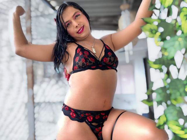 EmilyTsSweet with black and red lace lingerie, flexing arms, showing off her tits and pussy, smiling with red lipstick, black hair, and a gold necklace.