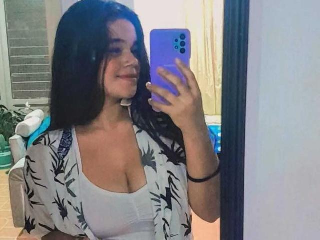 Victoria Boston, con cabello negro largo, un top blanco y una bata estampada, se toma un selfie frente al espejo con un teléfono morado, mostrando sus senos y sonriendo..