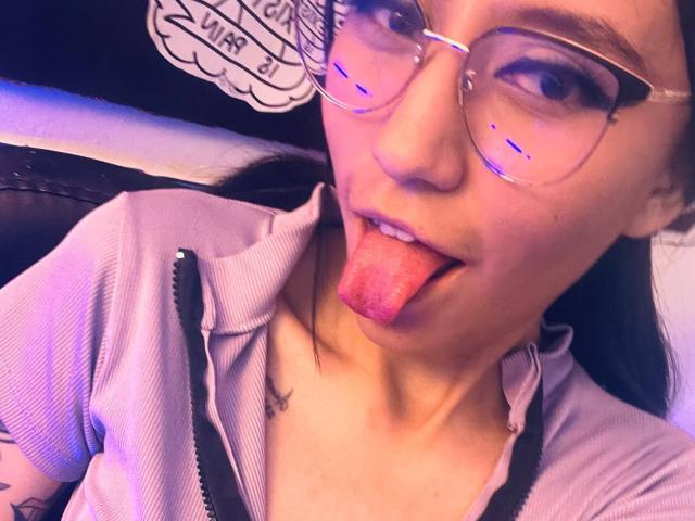 EviieGothel with glasses, tongue out, pink ribbed shirt, zipper partially down, visible tattoo on right arm, purple lighting, looking at camera, facial expression playful.