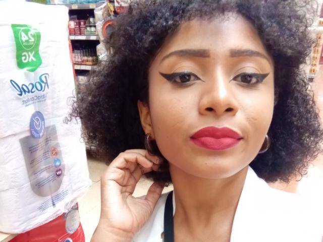 MeganWlliams has curly black hair, dark eyebrows, and red lipstick with dramatic black winged eyeliner. She's holding her hair with a white shirt on.