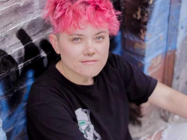 MeganC with short blonde hair, green eyes, nose ring, and black shirt, resting chin on hands, smiling slightly, on a bed with pink and gray pillows.