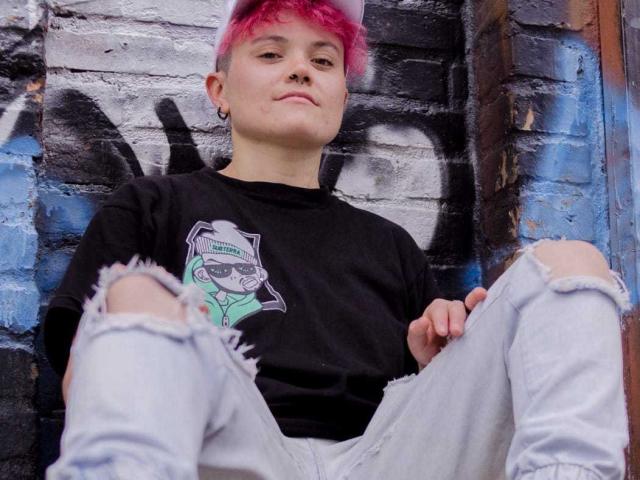 MeganC met roze krullend haar, een zwart T-shirt, een neusring en een lichte glimlach, zittend tegen een muur die bedekt is met graffiti. Ze heeft een bleke huid en blauwe ogen, en ze ziet er ontspannen uit..