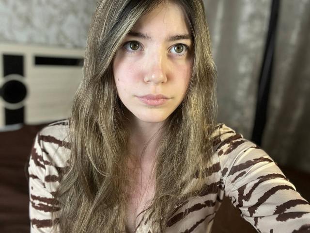 AmberSkylerr in lila lingerie, waarbij haar achterwerk in de focus staat, met één hand vastgehouden, tong uit, lang zwart haar, op een bed met roze en gestreepte kussens, in een kamer met roze verlichting..