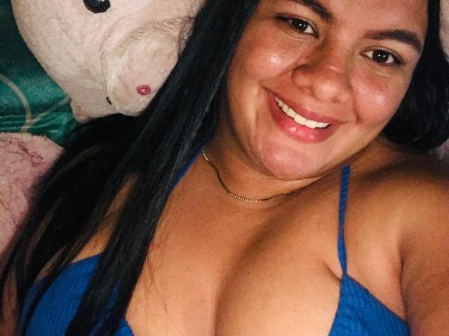 KloeLust in a white fishnet top, showing her tits, with a grey fur pillow, sitting on a bed, legs spread, revealing her pussy and tattoos, next to a keyboard.