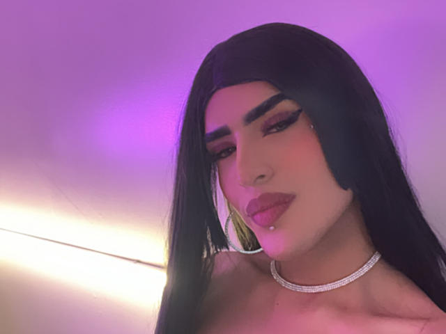 ScarlettTs with long black hair, purple-lit face, thick eyebrows, pink lipstick, pierced lip, silver choker, and a slight smile, showing off her pretty face and sexy look.