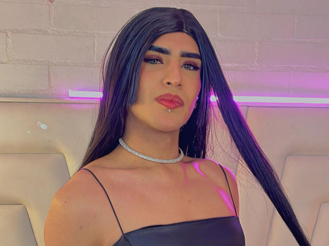 ScarlettTs with long black hair, thick eyebrows, red lipstick, and a lip piercing, wearing a black strap top and silver choker, has a sexy, confident look.