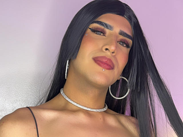 ScarlettTs has long black hair, thick eyebrows, bold makeup, red lips, a nose piercing, a chin piercing, large hoop earrings, and a sparkling choker, showing a confident, sexy look.