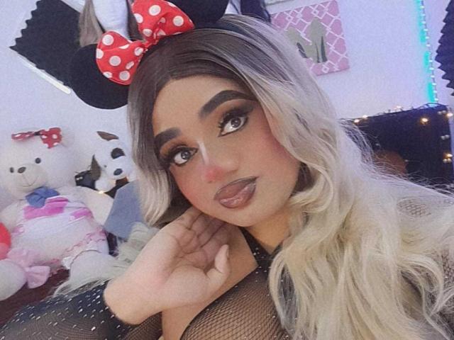 LunaSmithy: Eine Frau mit langen, blonden und grauen Haaren, die einen Minnie Maus-Haarband trägt, mit viel Make-up, einem Netzoberteil und vollen Lippen, die ihr Gesicht berührt. Ihre Brüste sind durch das Netzoberteil sichtbar..