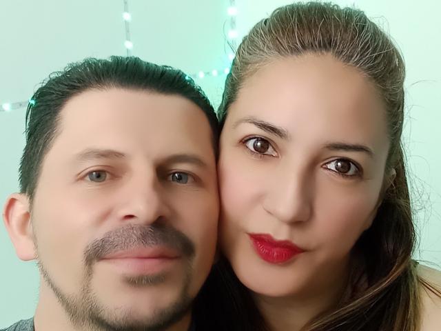 A KaelaX tem olhos castanhos, batom vermelho e cabelos longos castanhos, e está ao lado de um homem com bigode e cabelos pretos curtos. Ambos têm pele clara e estão sorrindo..