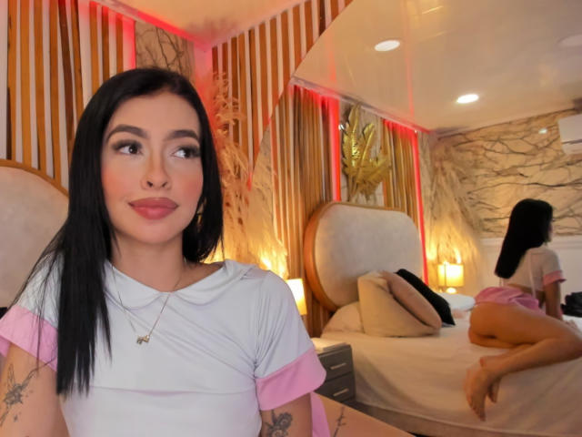 LeahMarie with black hair, pink and white shirt, tattoos, looking to the side, sitting in a room with another LeahMarie on a bed, pink skirt, and white top, facing away.