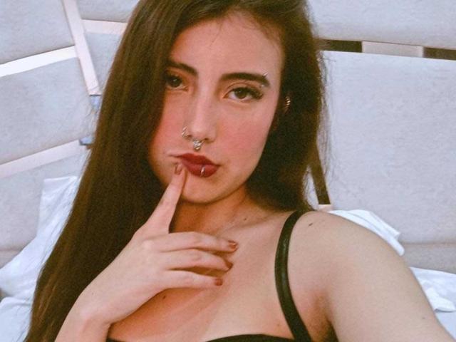 Diana Evans, con cabello castaño largo, un top negro con tirantes, un piercing en la nariz y un dedo en la boca, mostrando sus pechos, tiene una cara linda y está chupándose el dedo..