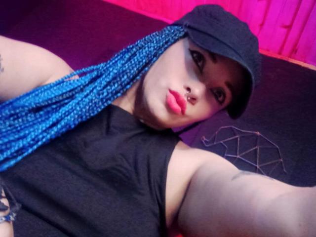 DalilaMusa with blue braids, black cap, black sleeveless top, nose ring, and pink lipstick, looking at camera, arms raised, showing tattoo on upper arm.