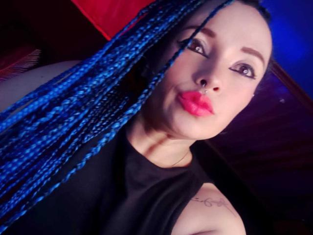 Dalila Musa, avec des tresses bleues, du rouge à lèvres, un anneau au nez, un haut noir sans manches, un tatouage sur l'épaule droite, regardant vers le haut, avec du khôl et du maquillage..