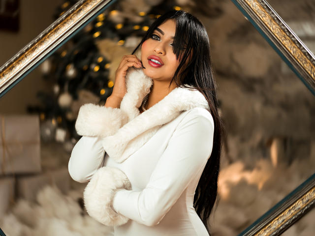 EmmaaRodriguez with long black hair, white fur-trimmed coat, red lipstick, hands near her face, smiling, looking at camera, Christmas tree with lights in blurred background.