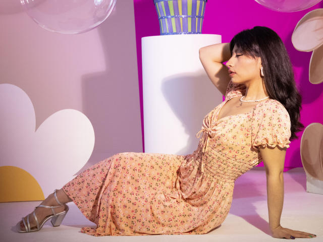 Selene Leon, con piel morena, cabello oscuro y pequeños senos, vestida con un vestido floral rosa, sentada con un brazo detrás de la cabeza, y tacones plateados, sobre un fondo colorido..