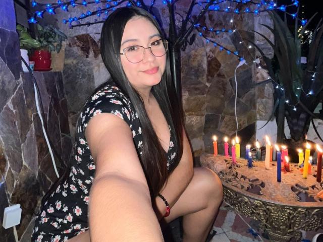 LyannaVega, with long black hair and glasses, is squatting, wearing a floral dress, showing her ass, and taking a selfie in front of lit candles and blue string lights.