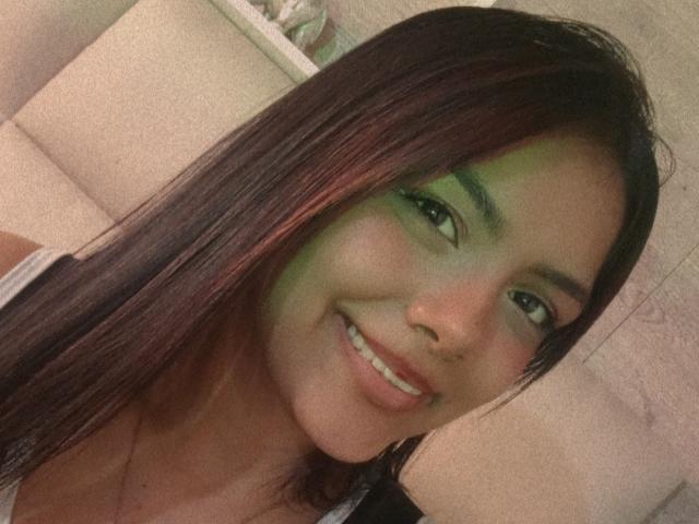 EmilyRoberts has dark brown hair with red highlights, green eye makeup, and is smiling with her teeth showing. Her ass is not visible. She's wearing a white top.