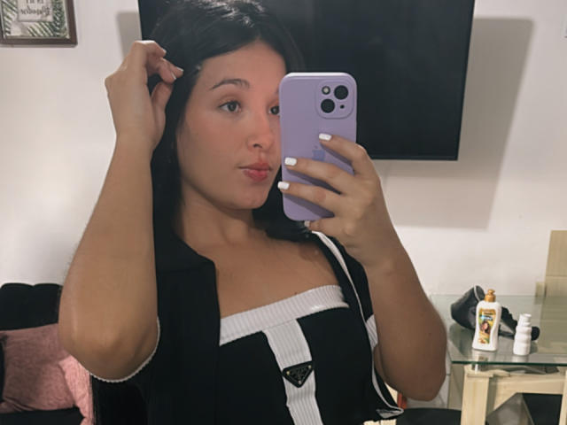 SarahGood with brown hair, pouty lips, and white nails, wearing a black vest over a white strap dress, taking a mirror selfie with a purple phone. Boobs visible.