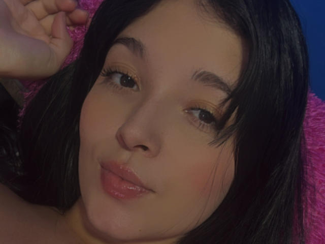SarahGood's face with glossy pink lips, dark straight hair, and golden eyeshadow, smiling slightly. Her right hand is partially visible, resting near her head.