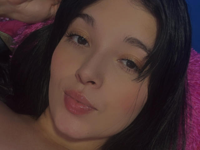 SarahGood's face with black hair, brown eyes, and glossy pink lips, looking slightly up with a subtle smile, her hand near her head. Her makeup includes yellow eyeshadow.