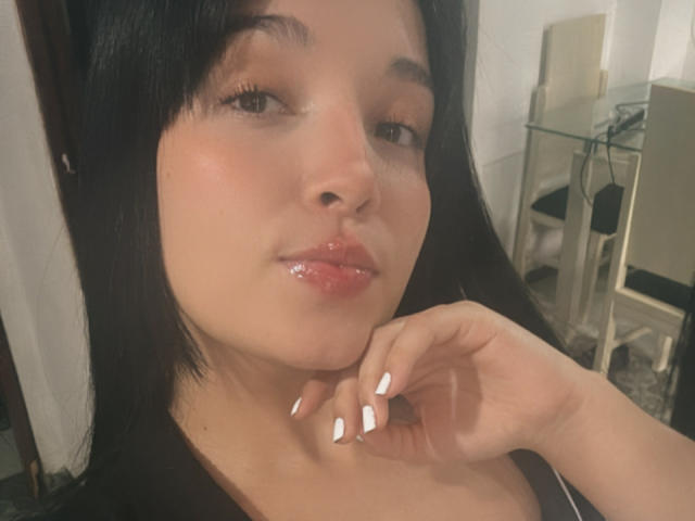 SarahGood with glossy pink lips, dark straight hair, white nail polish, and a slight smile, holding her chin. Her brown eyes are half-open, and she has a cute, soft look.