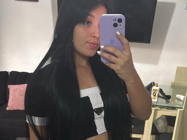 SarahGood with long black hair, white and black ribbed crop top, holding purple iPhone, taking mirror selfie, pouty lips, white nail polish, black and white shirt, medium tits.
