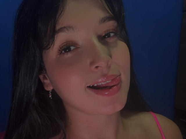 SarahGood with black hair, pink lipstick, and tongue out, has shiny wet lips, is smiling, and wearing pink earrings, against a dark blue background.